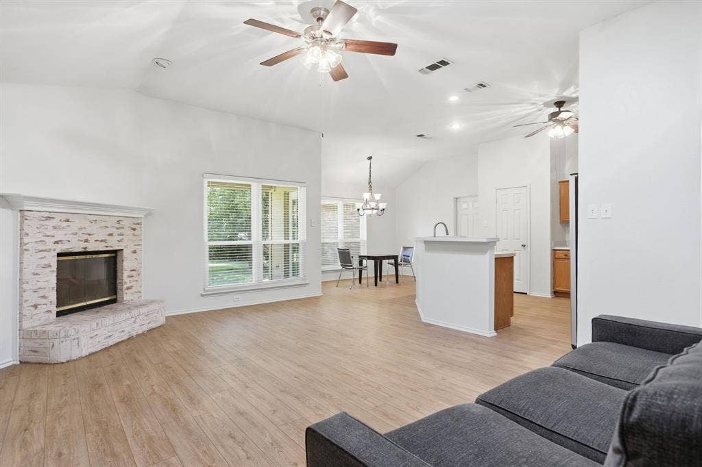 Open-concept living area featuring wood-finish flooring, a brick fireplace with a white mantel, and vaulted ceilings with ceiling fans