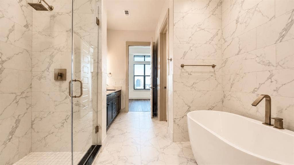 3330 Oak Grove Avenue, #1307, Dallas, TX 75204 Spacious bathroom featuring marble-look tile flooring and walls, a freestanding soaking tub with bronze-finish fixtures, and a frameless glass shower enclosure with a rain showerhead