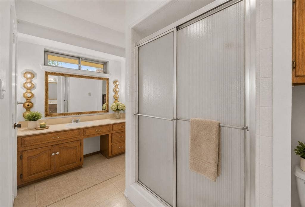 2125 Woodbrook Street, Denton, TX 76205 Shower enclosure with textured glass doors, a built-in wood-finish vanity with a light-toned countertop, a large framed mirror, and a transom window