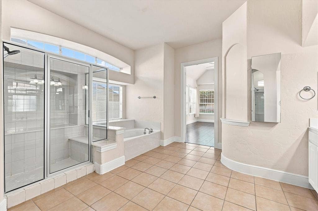 509 Wentworth Lane, Murphy, TX 75094 Spa-inspired primary bathroom featuring a frameless glass-enclosed shower with tile surround, soaking tub with deck-mounted fixtures, dual vanity with medicine cabinet, tile flooring, and abundant natural light from transom windows