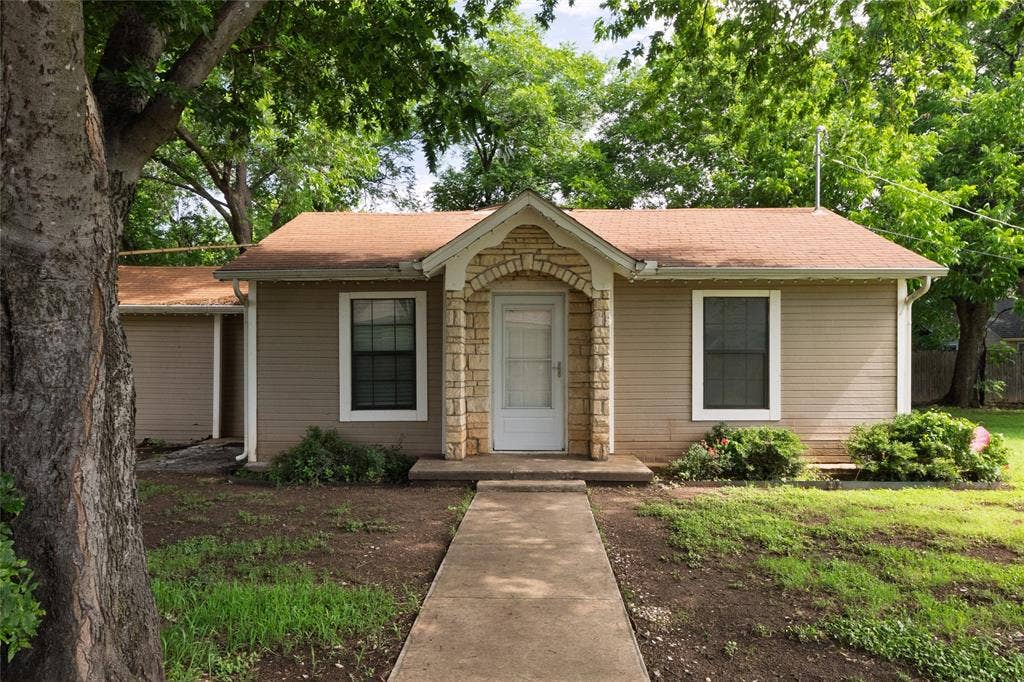 313 N Roe Street, White Settlement, TX 76108 Front exterior featuring a stone archway entryway, two windows with white trim, tan siding, and a brown shingle roof