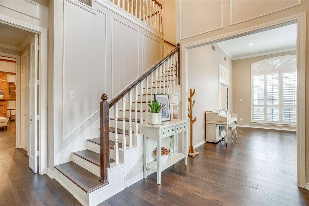 4624 Reunion Drive, Plano, TX 75024 Foyer featuring a grand staircase with white risers and dark wood treads, extensive wall paneling, and a wide entryway leading to a formal living room with a large arched window with plantation shutters