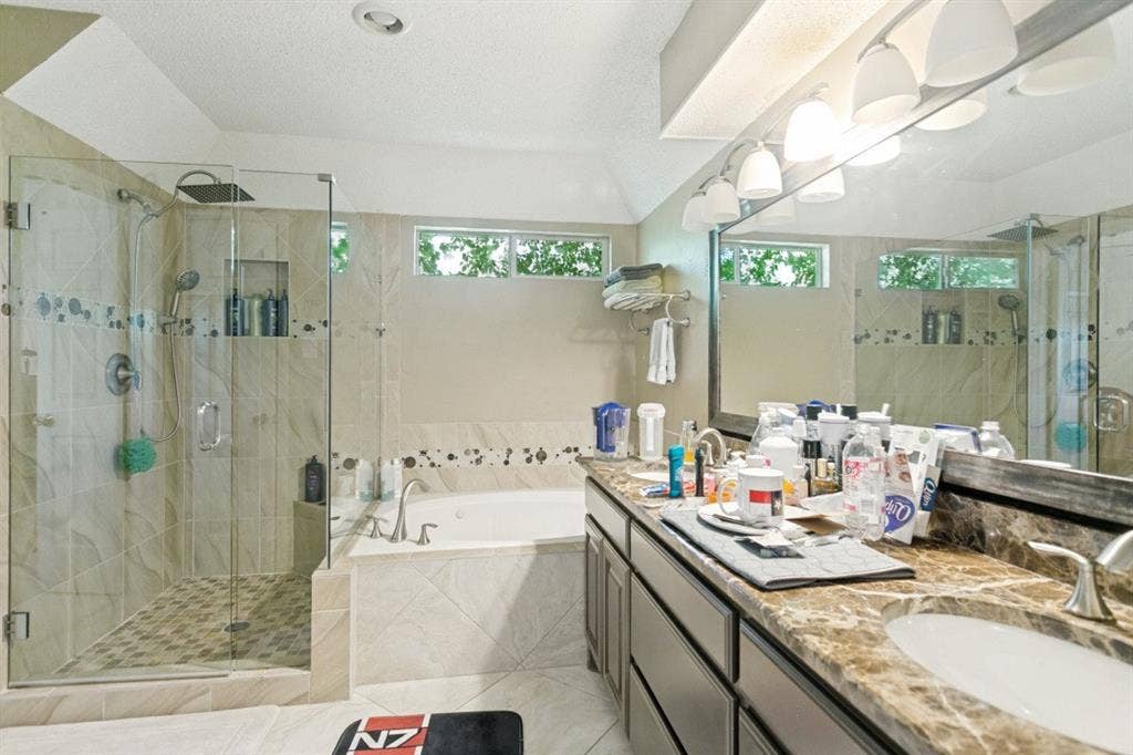 6713 Ashglen Place, Plano, TX 75023 Spacious bathroom featuring a glass-enclosed walk-in shower with a rain showerhead, a separate built-in soaking tub, a double vanity with an integrated mirror, warm-toned countertops, and multiple transom windows