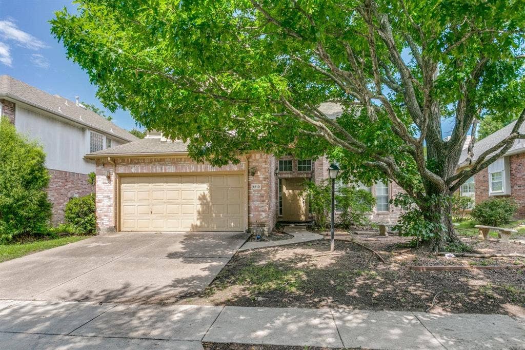 6713 Ashglen Place, Plano, TX 75023 Brick facade residence with an attached garage, featuring a mature tree, concrete driveway, and front sidewalk