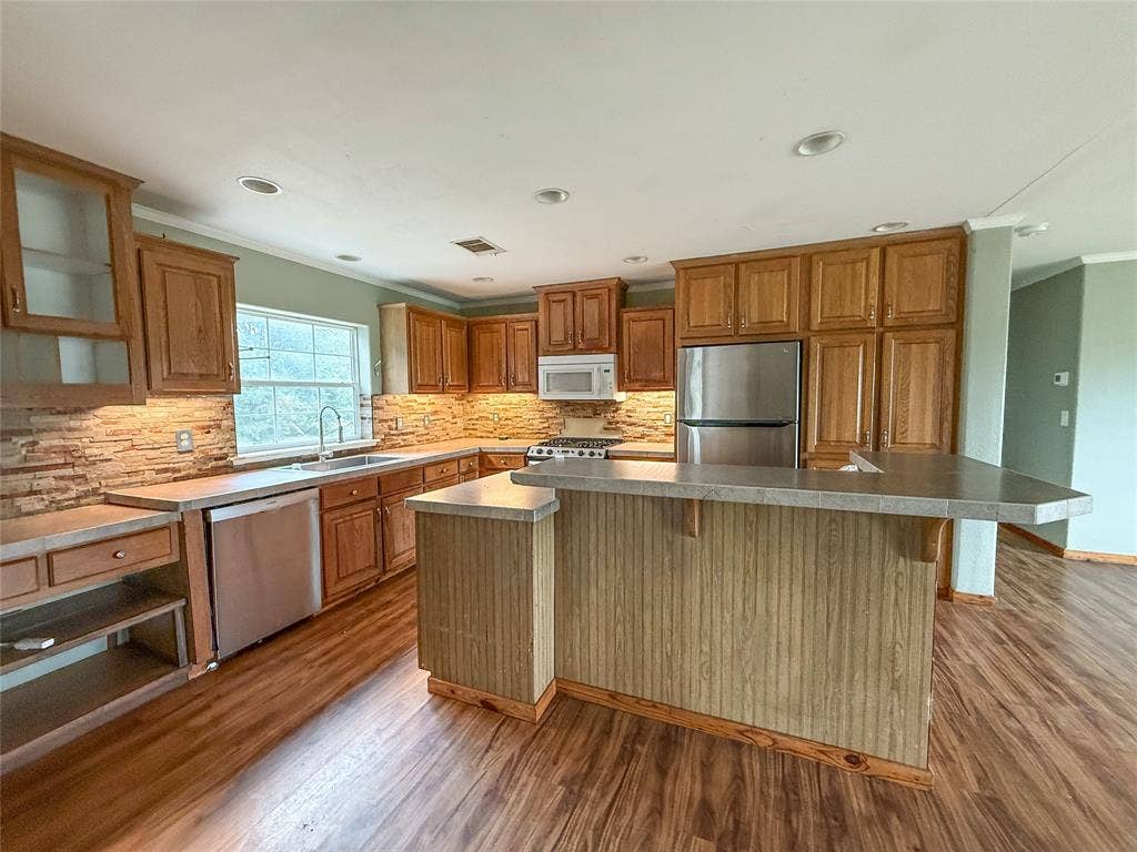 2749 Mcgregor South Loop, McGregor, TX 76657 Kitchen featuring wood-finish cabinetry, stainless steel appliances, a stone tile backsplash, recessed lighting, and an island with a breakfast bar