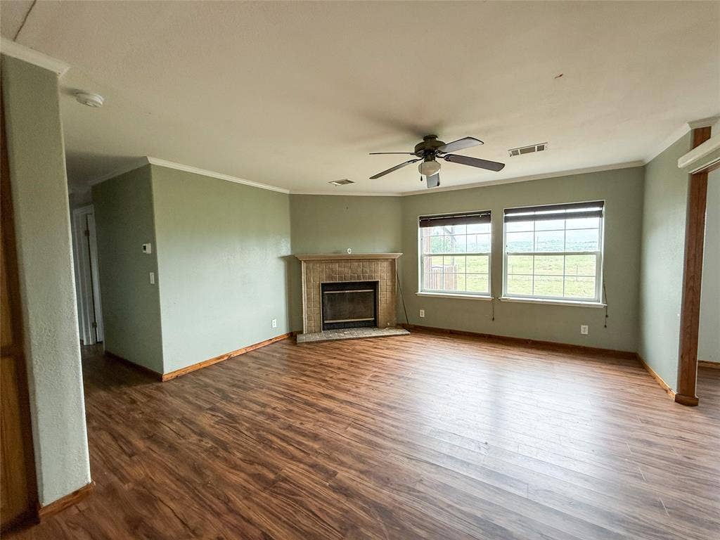 2749 Mcgregor South Loop, McGregor, TX 76657 Spacious living area featuring wood-finish flooring, a fireplace with a tiled surround and wood mantel, two large windows, a ceiling fan, and crown molding