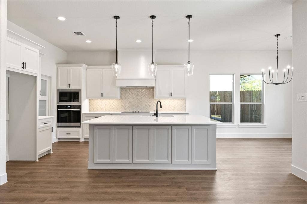902 Boston Lane, River Oaks, TX 76114 Open-concept kitchen featuring an oversized island with a built-in sink, white shaker cabinetry, herringbone tile backsplash, integrated double ovens, and wood-finish flooring