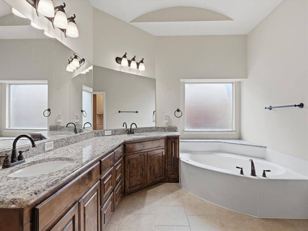 1621 Lakemere Drive, Prosper, TX 75078 Bathroom featuring a corner soaking tub, dual vanities with granite countertops, and wood-finish cabinetry