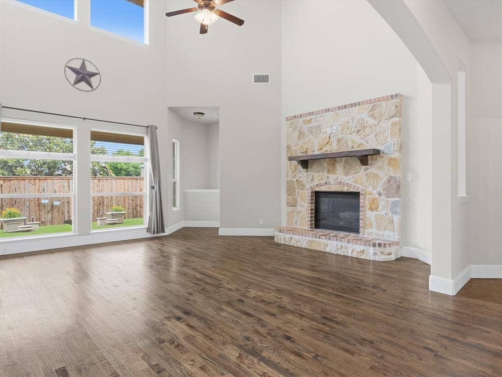 1621 Lakemere Drive, Prosper, TX 75078 Spacious living area featuring high ceilings, wood-finish flooring, a stone fireplace with a substantial wood mantel, a ceiling fan, and large windows offering a view of the fenced backyard
