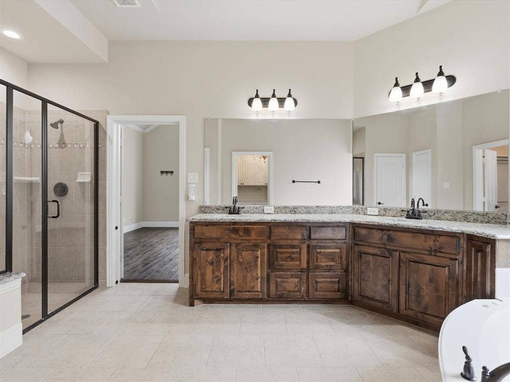 1621 Lakemere Drive, Prosper, TX 75078 Spacious bathroom featuring a frameless glass shower enclosure, dual sink vanity with granite countertops, rich wood cabinetry, and neutral tile flooring
