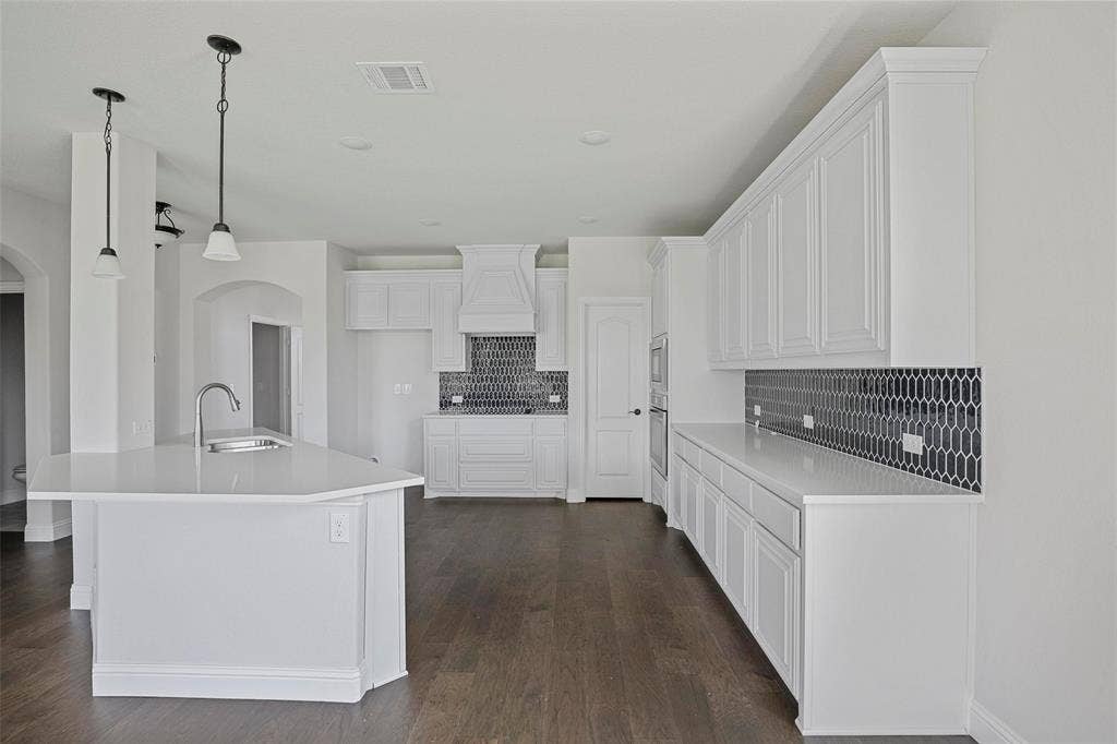 508 Bush Avenue, Waxahachie, TX 75165 Bright kitchen featuring white cabinetry, a large central island with an undermount sink, dark wood-finish flooring, a patterned tile backsplash, and stainless steel built-in appliances