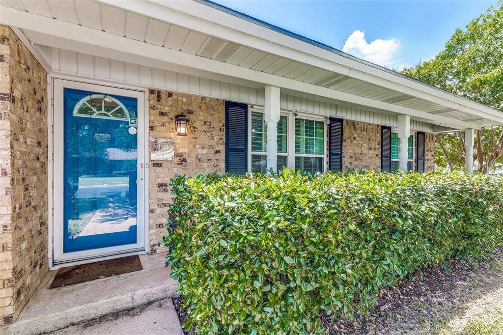 1121 W Redbud Drive, Hurst, TX 76053 Brick exterior featuring a storm door with an arched transom, a covered porch with white columns, windows with dark louvered shutters, and a manicured hedge