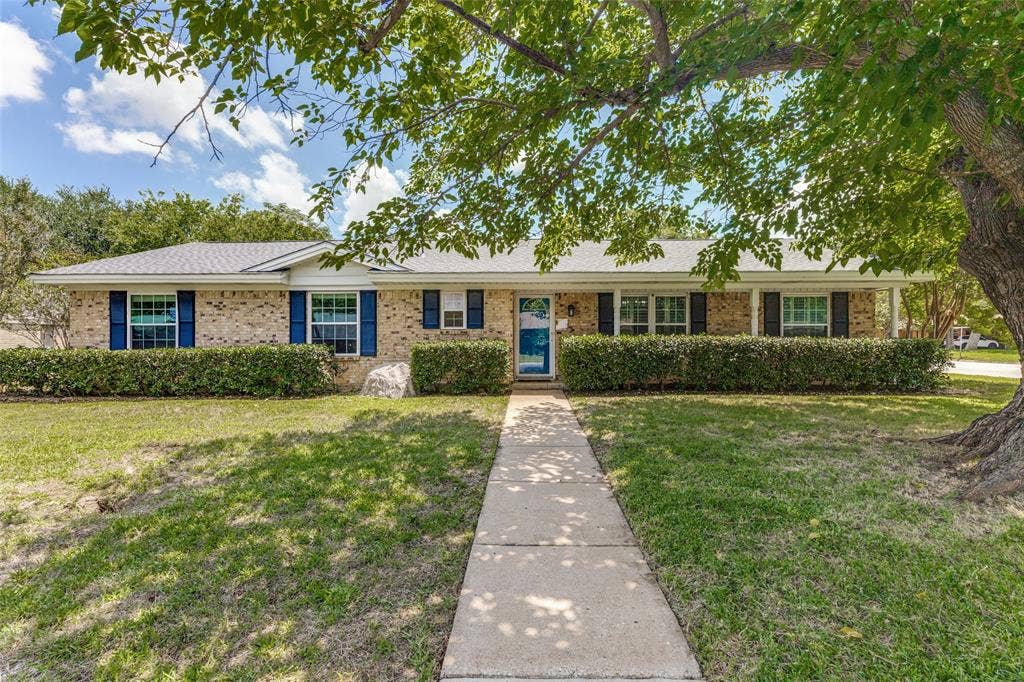 1121 W Redbud Drive, Hurst, TX 76053 Brick facade ranch-style residence featuring a white roofline, black window shutters, and a solid blue entry door