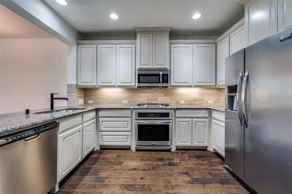 7424 Mitchell Drive, McKinney, TX 75070 Gourmet style kitchen in McKinney Texas townhome featuring stainless steel appliances, light stone countertops, dark wood type flooring, white cabinets, recessed lighting, and tile backsplash, delivering the updated kitchen features and clean modern look today’s Collin County buyers search for when shopping online for McKinney TX townhomes for sale with upgraded kitchens.