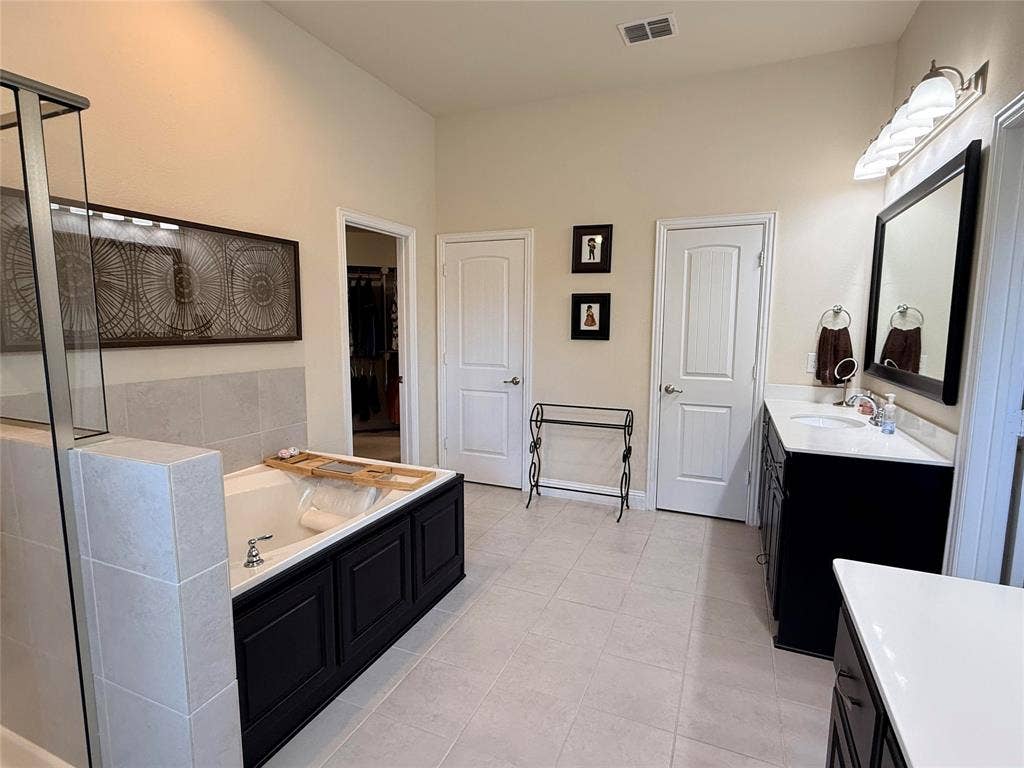7310 Venice Drive, #4, Grand Prairie, TX 75054 Spacious bathroom featuring a built-in soaking tub with black paneled surround, a separate glass-enclosed shower, a double vanity with dark cabinetry and light-colored countertops, and light-toned tile flooring