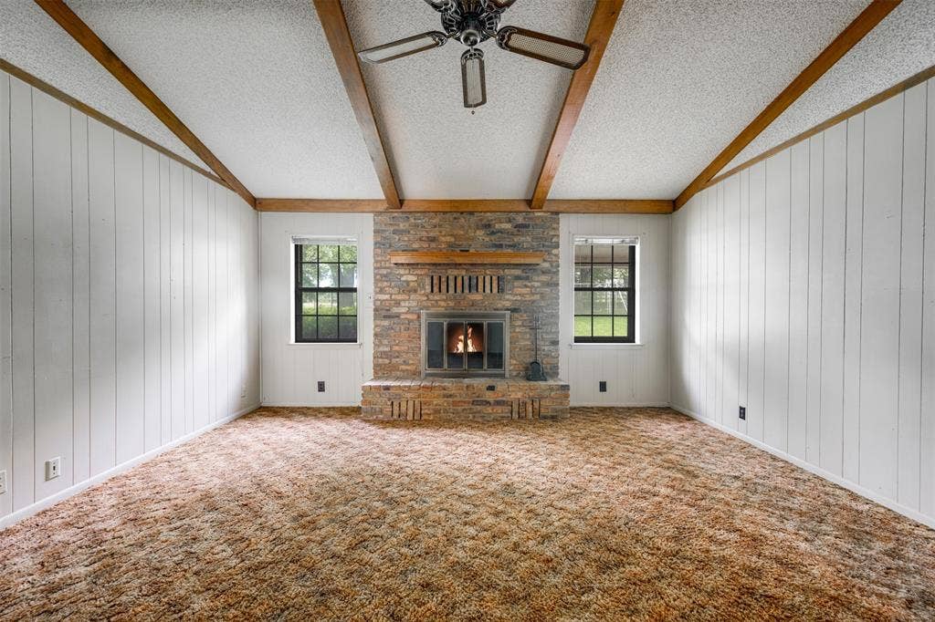 8798 Hill N Dale Road, Tyler, TX 75709 Spacious room featuring a brick fireplace with a wood mantle, exposed wood ceiling beams, white vertical paneling, and two windows with black frames