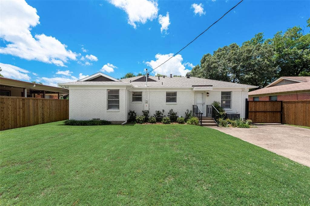 2523 San Medina Avenue, Dallas, TX 75228 Expansive backyard featuring a lush lawn, white brick exterior, mature perimeter trees, wood privacy fencing, and a concrete driveway