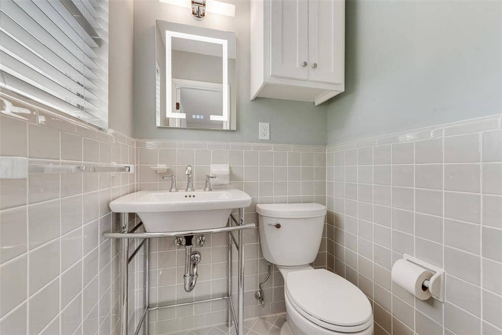 2523 San Medina Avenue, Dallas, TX 75228 Bathroom featuring light grey square wall tiling, a pedestal sink with chrome stand, a wall-mounted lighted vanity mirror, and a white storage cabinet