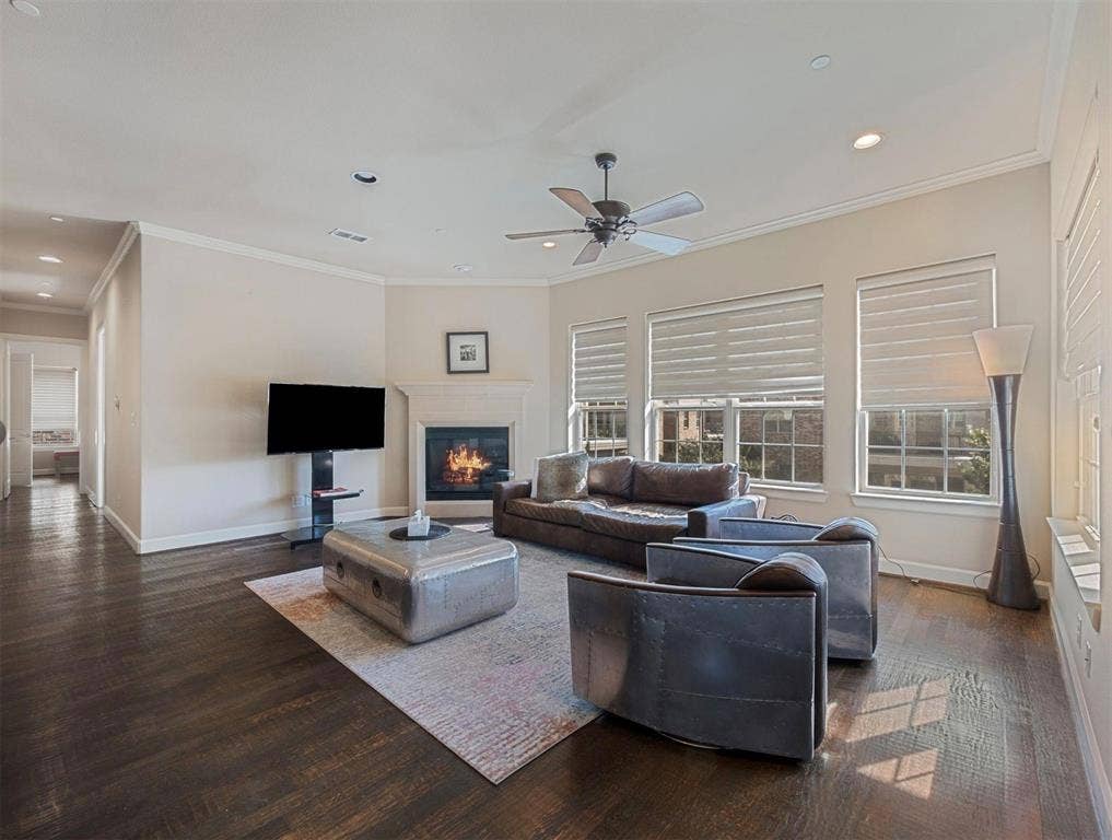 5001 Meridian Lane, #2103, Addison, TX 75001 Spacious living area featuring dark wood-finish flooring, a corner fireplace with a mantel, multiple windows with blinds, crown molding, and recessed lighting