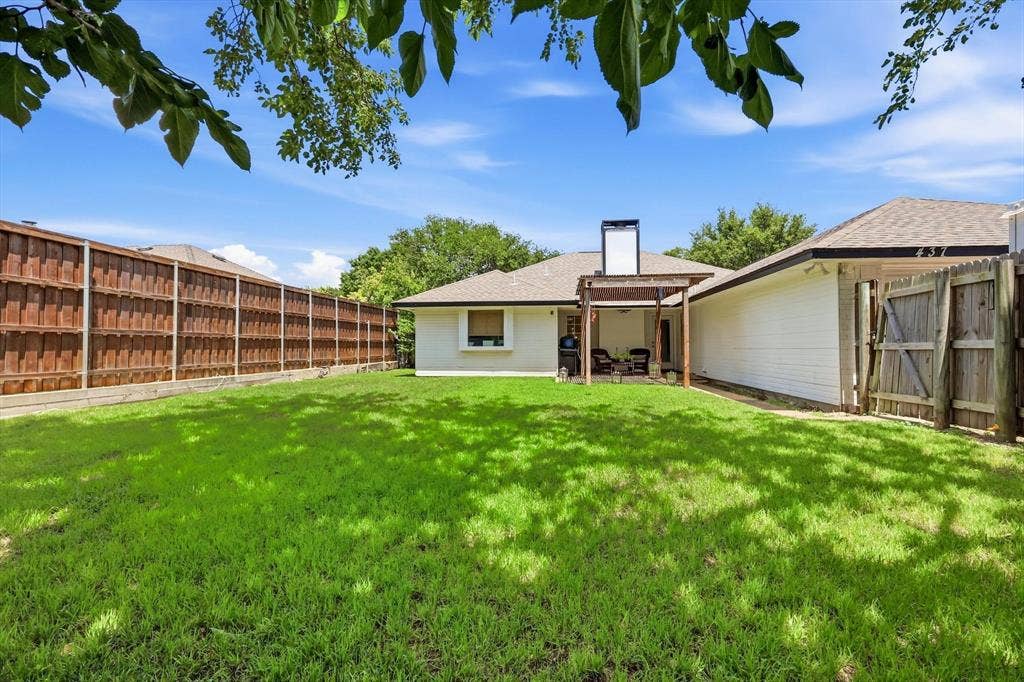 437 Bradshaw Street, Cedar Hill, TX 75104 Expansive backyard featuring a lush lawn, a covered patio with a wooden pergola, a white-painted exterior, a perimeter wooden fence, and a gate providing side yard access