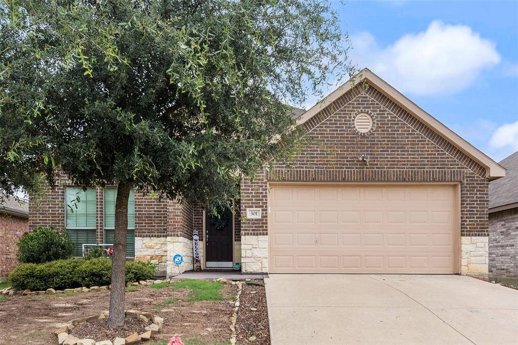 301 Billy Wickliffe Drive, Wilmer, TX 75172 Brick and stone facade residence featuring a two-car garage, mature front yard tree, concrete driveway, and front entryway with a dark-paneled door
