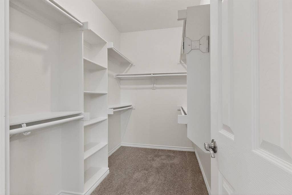 217 Gatehouse Drive, Red Oak, TX 75154 Walk-in closet featuring built-in shelving, white painted wood rods, a white paneled door with a brushed nickel handle, and carpet flooring
