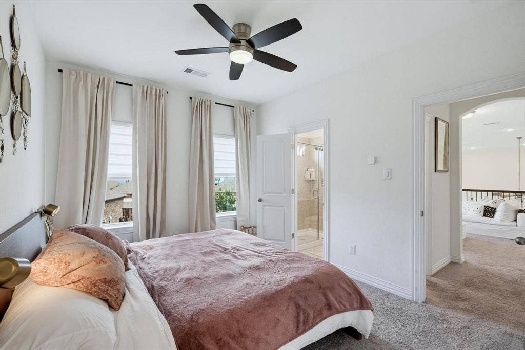 1205 Shady Fox Way, Arlington, TX 76005 Carpeted bedroom featuring three windows with blinds and drapes, a ceiling fan with integrated lighting, an arched doorway, and a white paneled door revealing a tiled shower with a glass enclosure