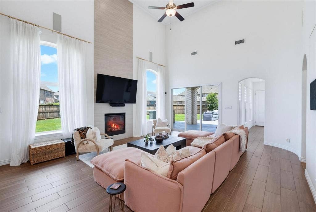 1205 Shady Fox Way, Arlington, TX 76005 Spacious living area featuring a two-story ceiling, wood-finish flooring, a contemporary fireplace with a mounted television above, and large arched windows with sheer curtains