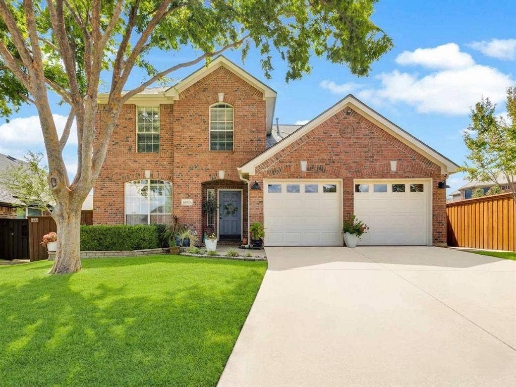 6004 Crestmill Lane, Sachse, TX 75048 Brick facade residence featuring a two-car garage with panel windows, a concrete driveway, a manicured lawn, and mature trees