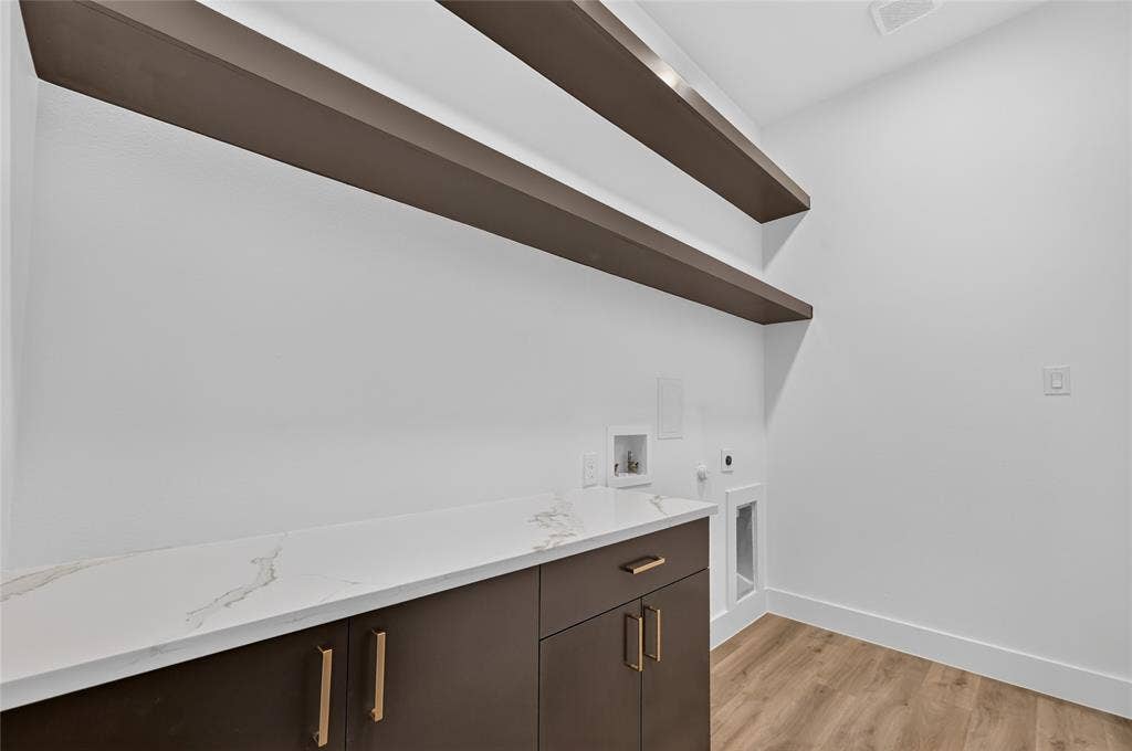 5130 Echo Avenue, Dallas, TX 75215 Laundry room featuring a white countertop with subtle veining, dark brown cabinetry with gold hardware, and two long floating shelves