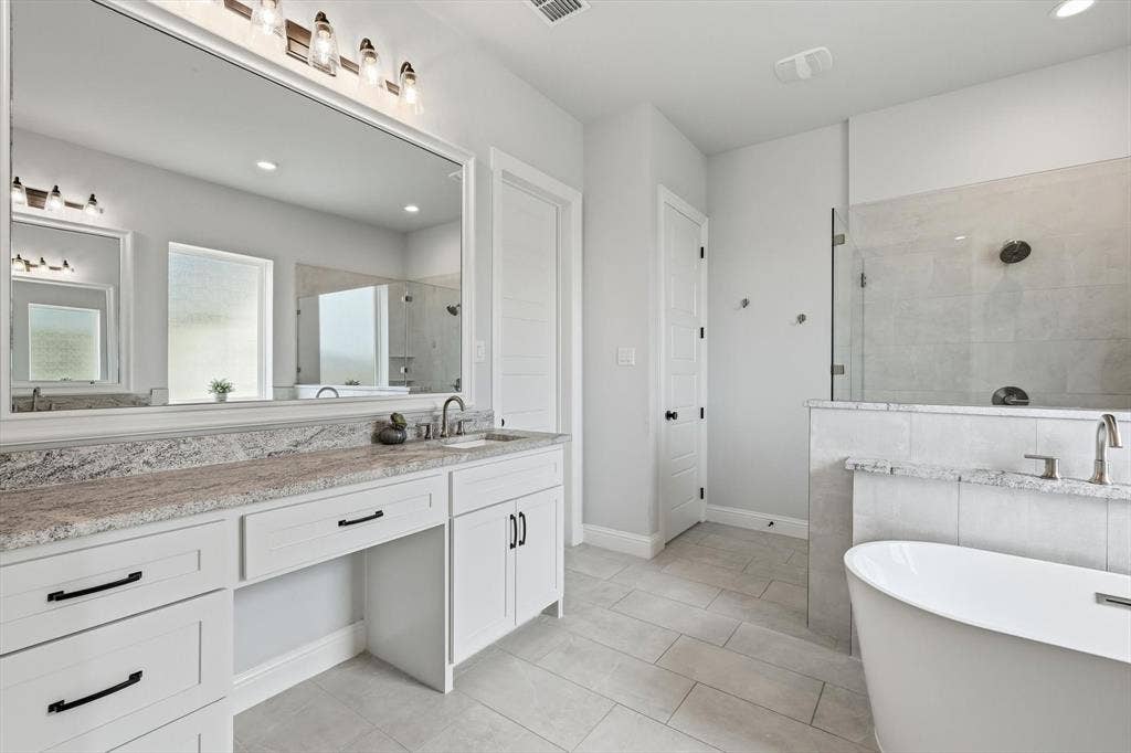 Spacious bathroom featuring a dual vanity with stone countertops, a large frameless mirror, a freestanding soaking tub, a separate walk-in shower with a glass enclosure, and light-toned tile flooring