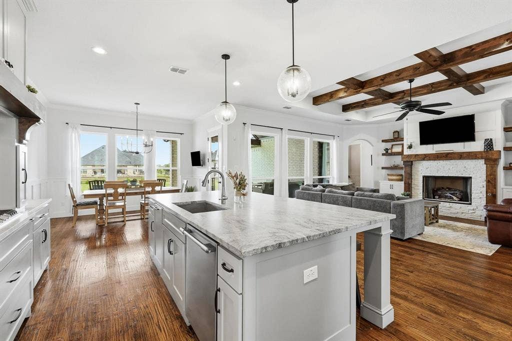Spacious kitchen island with a built-in sink and dishwasher, wood-finish flooring, a dining area with ample natural light, and an adjacent living area featuring a fireplace with a white stone surround and wood mantel, complemented by exposed ceiling beams