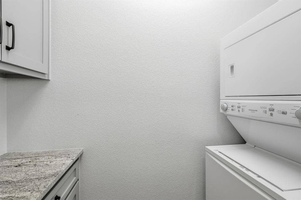 Guest Home - Laundry area featuring a stacked washer and dryer, light gray cabinetry with black hardware, and a stone-finish countertop