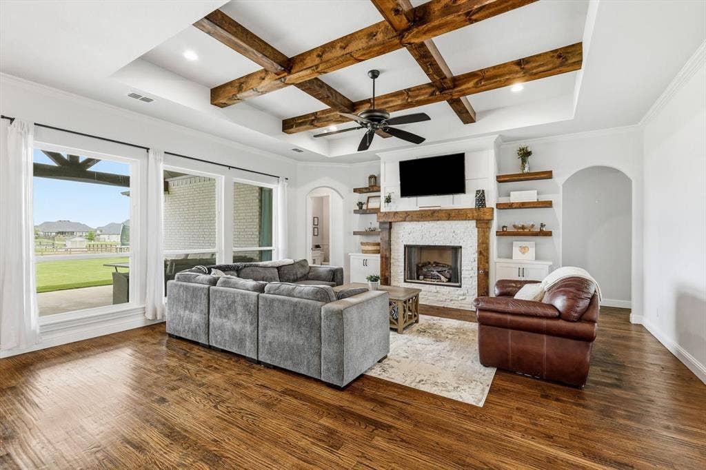 Spacious living area featuring exposed wood ceiling beams, a white brick fireplace with a wood mantel, built-in shelving, wood-finish flooring, and large windows