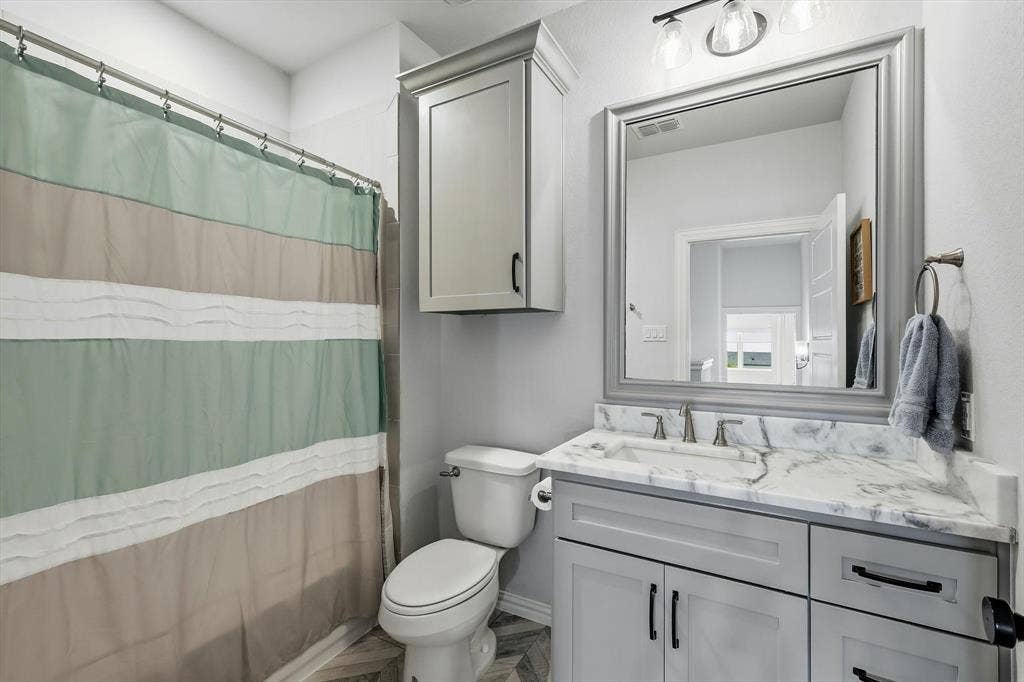 Upstairs bathroom featuring a single vanity with a patterned countertop, gray cabinetry with dark hardware, a framed mirror, and a wall-mounted light fixture