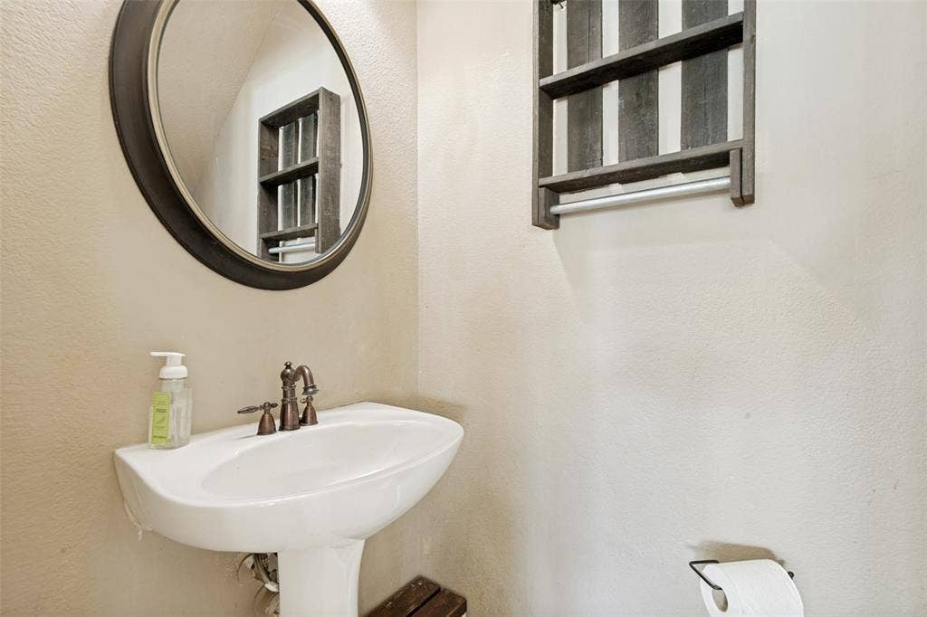 605 Rock Brook Drive, Forney, TX 75126 Pedestal sink featuring a bronze-finish faucet, circular framed mirror, and a wall-mounted wooden shelf unit with a towel bar