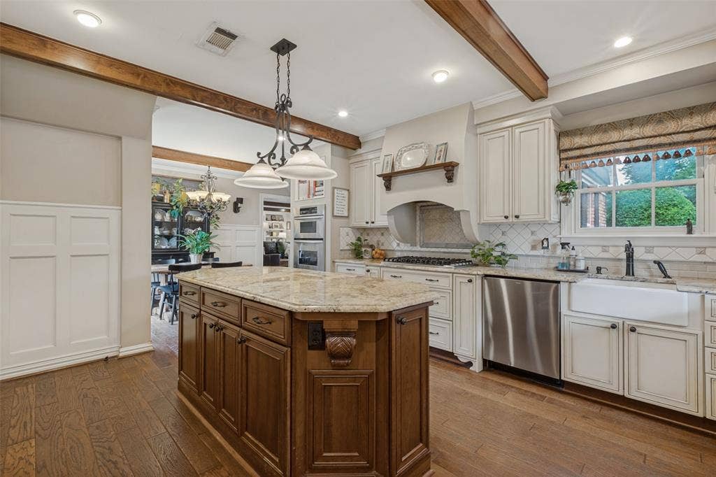 Kitchen featuring a wood-tone center island, stone countertops, farmhouse sink, exposed ceiling beams, and built-in double wall ovens