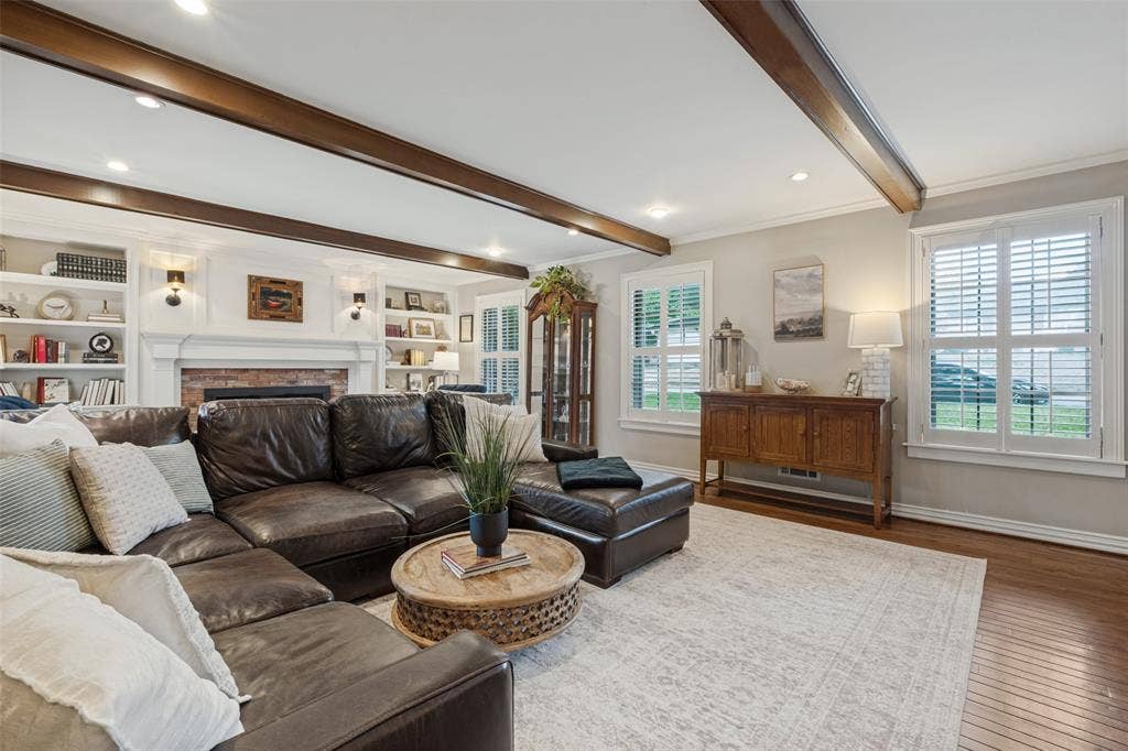 Living area featuring dark stained exposed ceiling beams, wood-finish flooring, built-in floor-to-ceiling bookshelves, and a brick fireplace surround