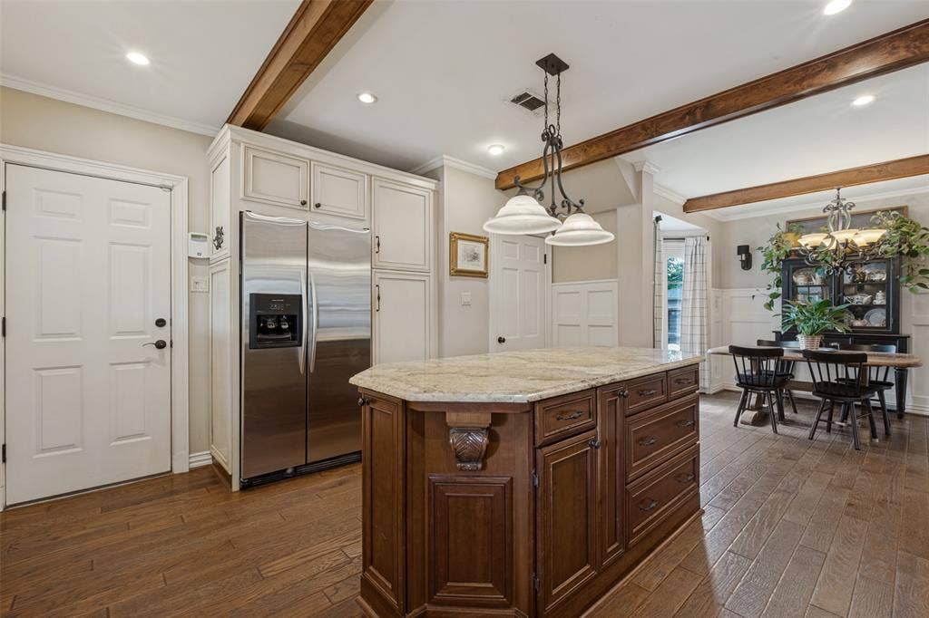 Kitchen featuring wood-finish flooring, exposed stained-wood ceiling beams, a central island with stone countertop, and custom cabinetry with stainless steel refrigerator