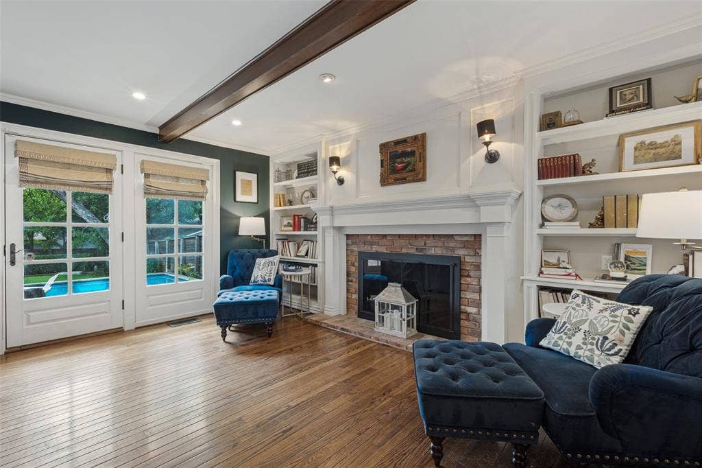 Wood-finish flooring, fireplace with a brick surround, built-in floor-to-ceiling shelving, exposed ceiling beam, and French doors