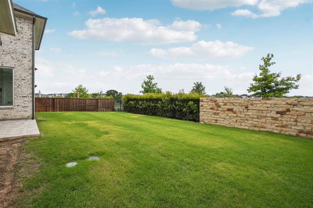 3960 Medio Creek Drive, Prosper, TX 75078 Expansive backyard featuring a manicured lawn, a full privacy fence, a stone retaining wall, and a tiled patio area