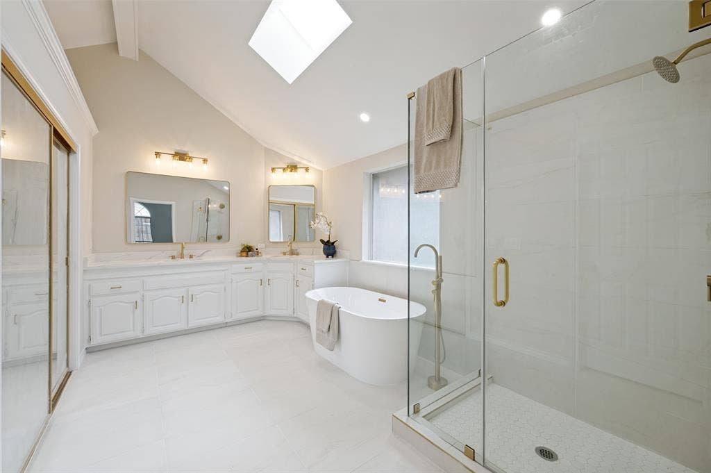 4420 Barwyn Lane, Plano, TX 75093 Vaulted ceiling bathroom featuring a large skylight, dual vanity with white cabinetry, freestanding soaking tub, and a glass-enclosed shower with gold-tone fixtures