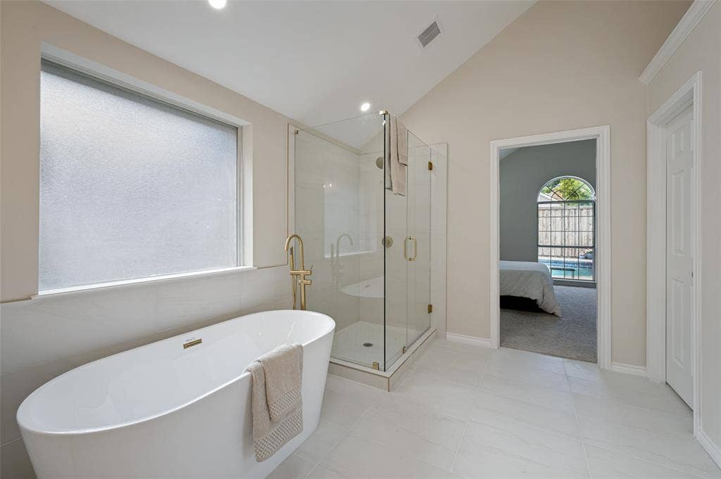 4420 Barwyn Lane, Plano, TX 75093 Spacious bathroom featuring a freestanding soaking tub with brass fixtures, a glass-enclosed shower, vaulted ceilings, frosted window, and large format floor tiling