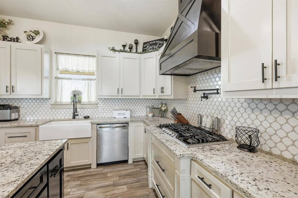 Kitchen featuring white shaker-style cabinetry, patterned tile backsplash, light-toned stone countertops, a farmhouse sink, and wood-finish flooring