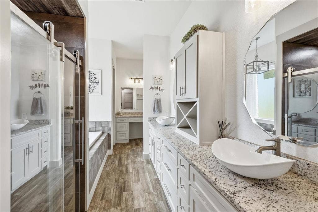 Dual vessel sinks set on a light granite-finish countertop, expansive vanity cabinetry, wood-look flooring, and a large soaking tub with a tiled surround