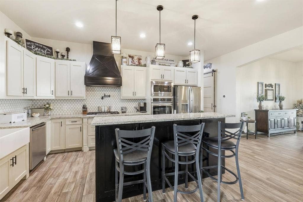 Kitchen featuring a large island with counter seating, a white apron-front sink, stainless steel appliances, a black range hood, and wood-finish flooring