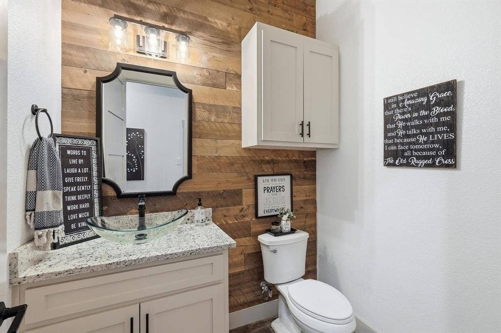 Wood-finish accent wall framing a vanity with a granite-style countertop, glass vessel sink, and an ornamental mirror