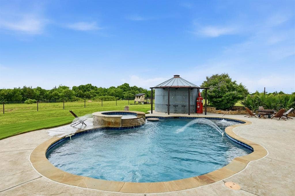 Curved swimming pool with integrated spa and water features, surrounded by a stamped concrete patio with stone coping