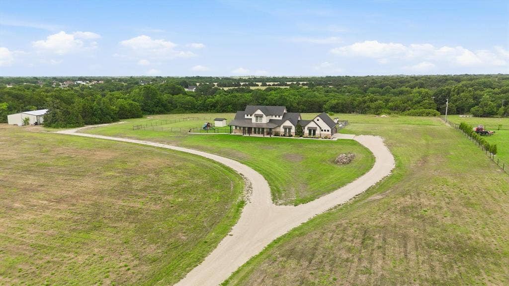 Spacious rural property featuring a large residence with an expansive covered front porch, a separate metal-roofed outbuilding, and a winding gravel driveway