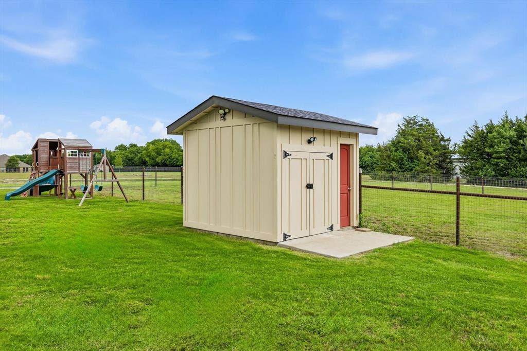 Detached utility shed featuring board and batten siding, a gray shingle roof, exterior lighting, and a concrete pad